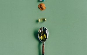 A green background with a silver teaspoon holding liquid. Some pills and tumeric in piles line up with the spoon.